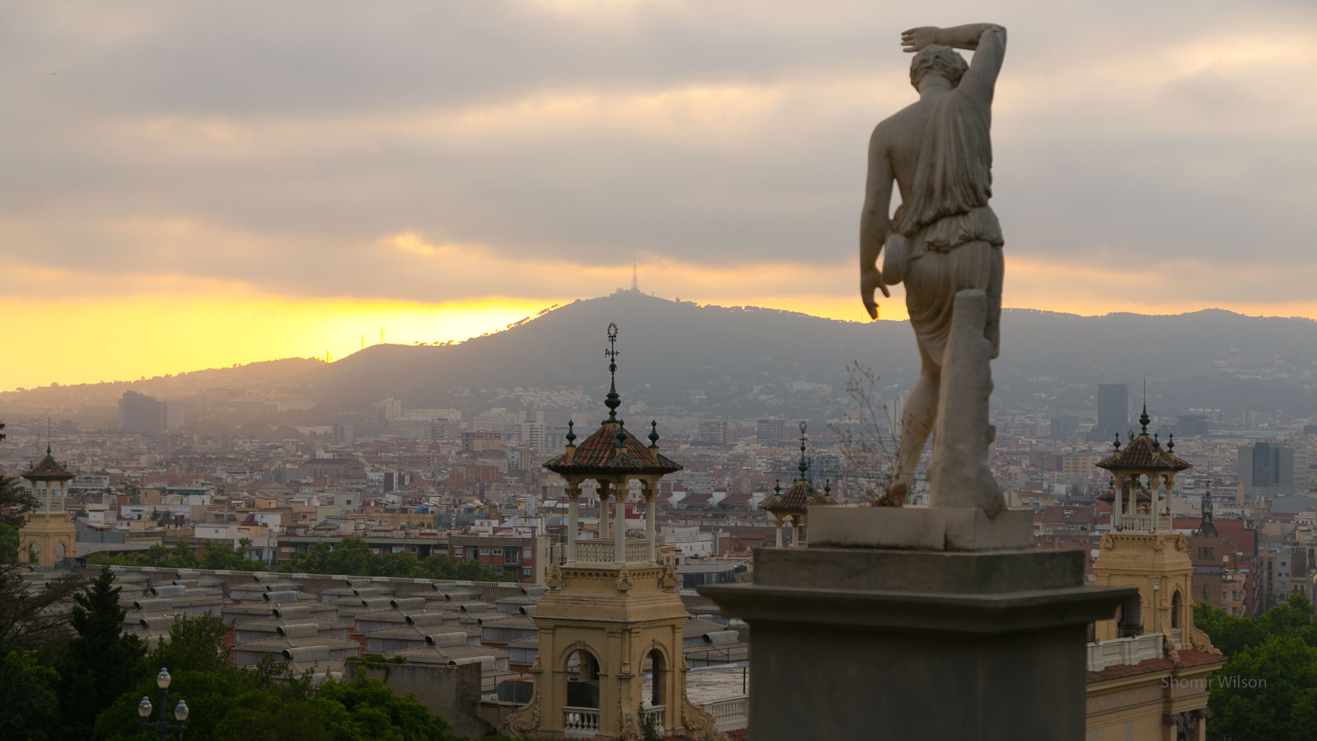Catalan city skyline from a hillside, with a statue of a person at right, and sunset in the sky