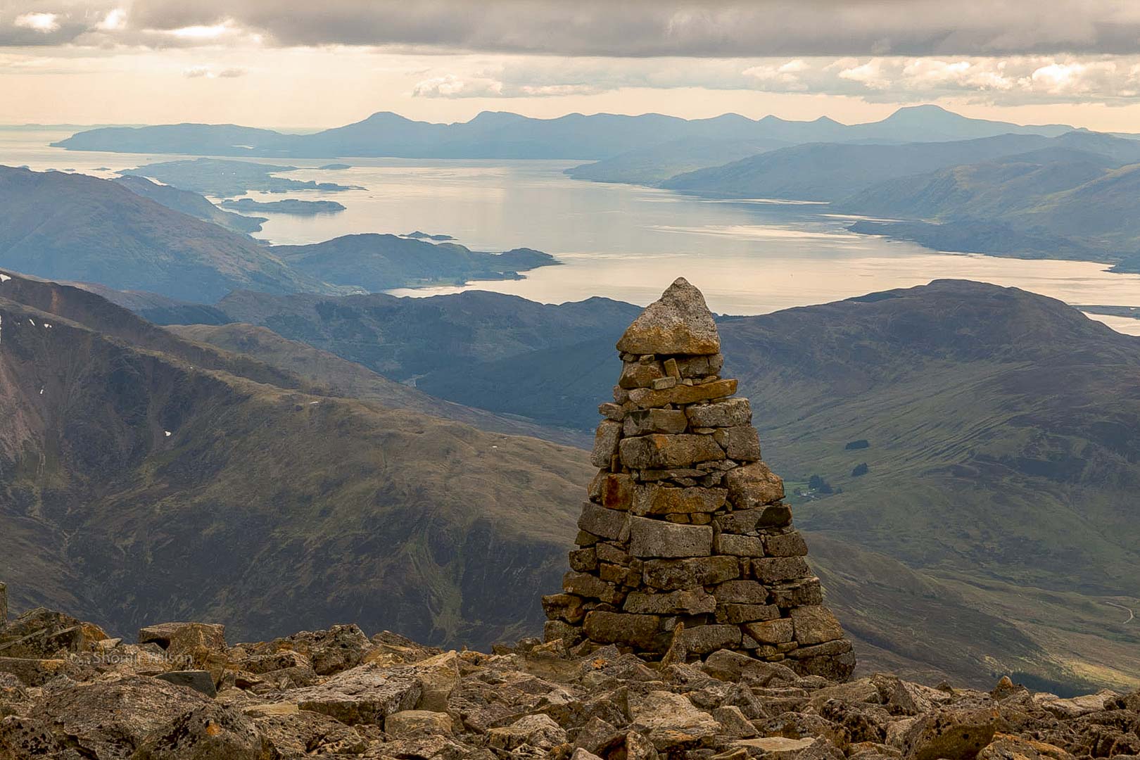 at a high altitude, a cairn of stones surrounded by stones on the ground, with mountains and a sea inlet behind