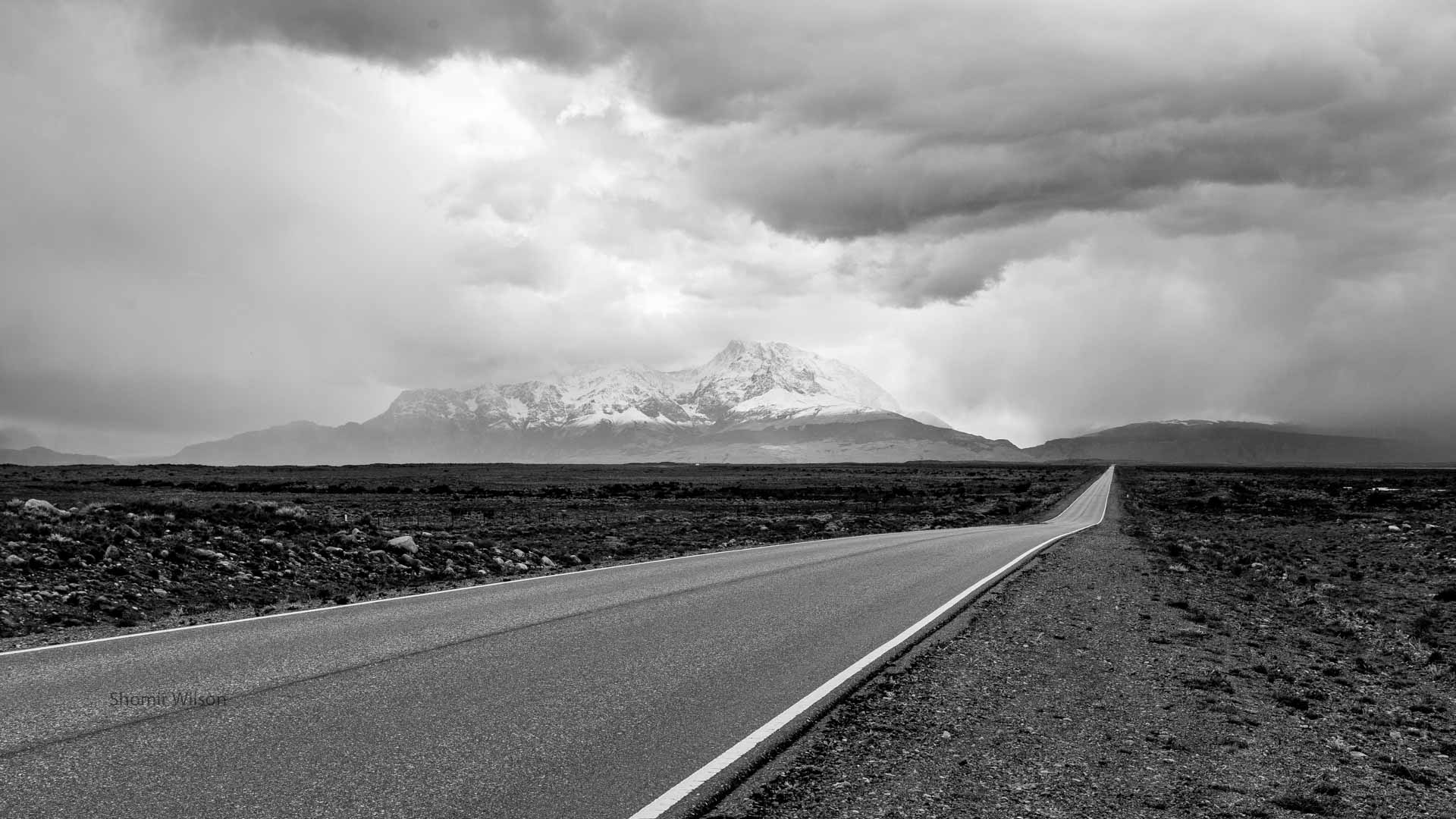 in monochrome: a two-lane road straight into the far distance across a rocky desert, with snow-capped mountains in the distance and heavy clouds above