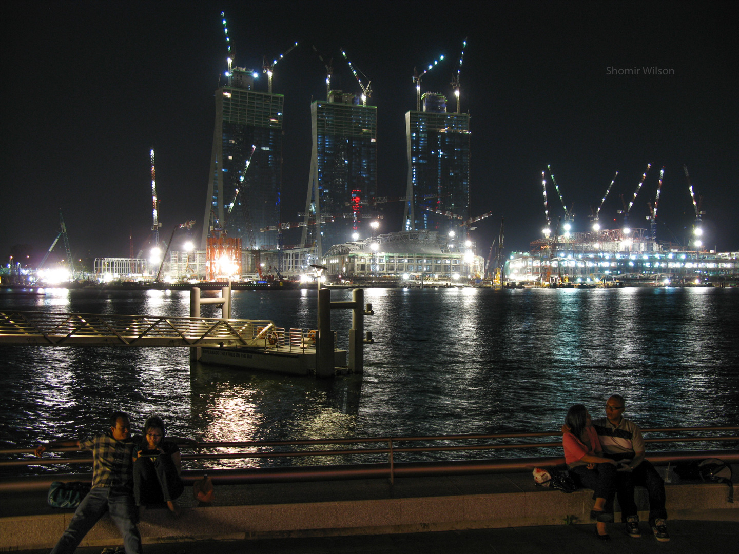 Brightly lit construction site across the water from the foreground, in which two couples canoodle