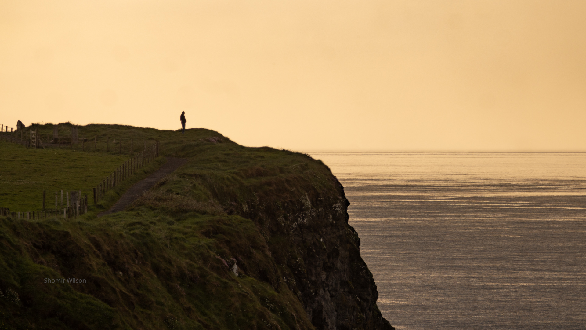 slihouette of a person on a tall cliff with the sea in the background; gentle orange sunset colors