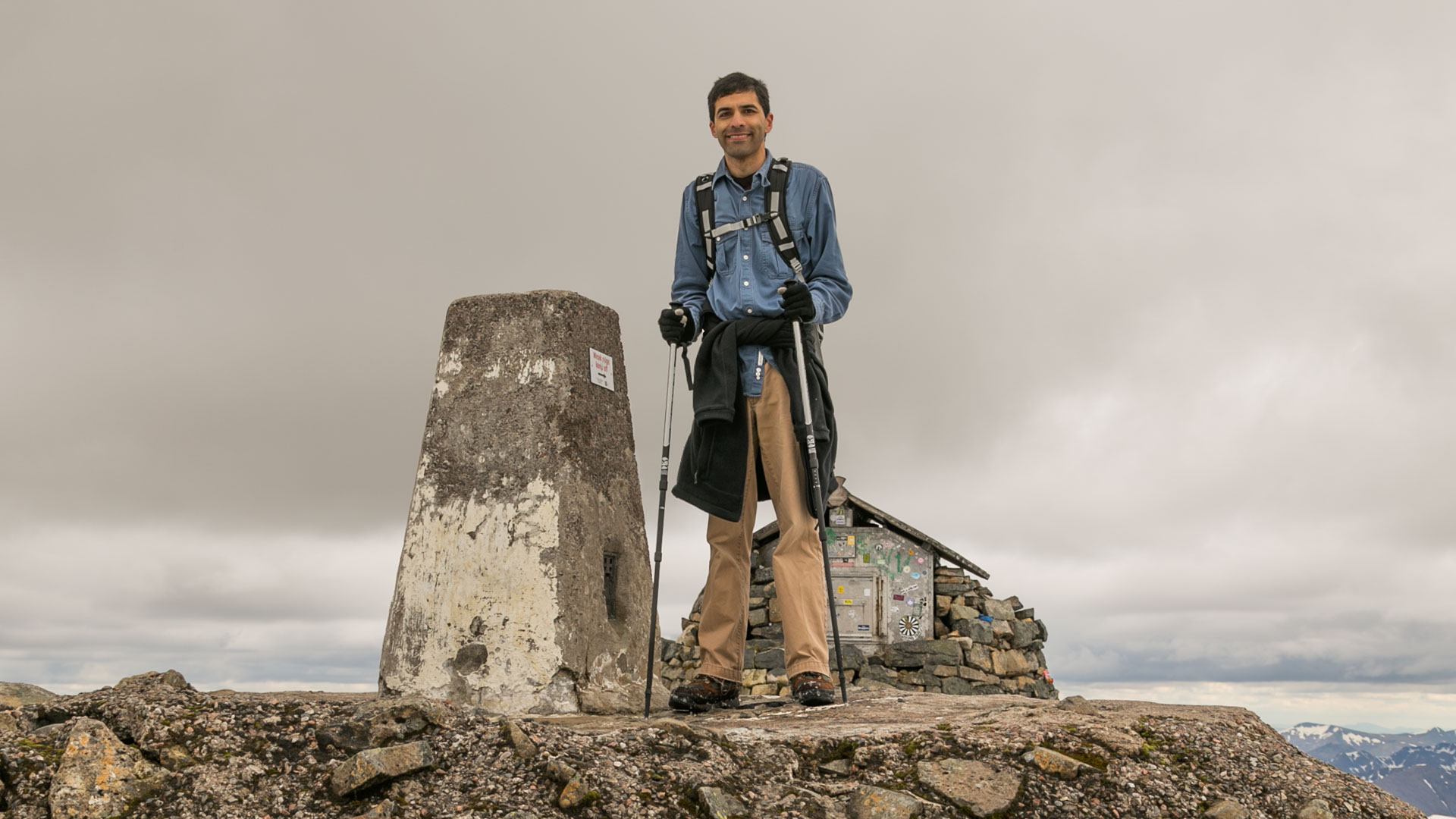Me with trekking poles, standing next to a cairn on top a rocky platform, with a metal shack in the background and a gray sky behind.