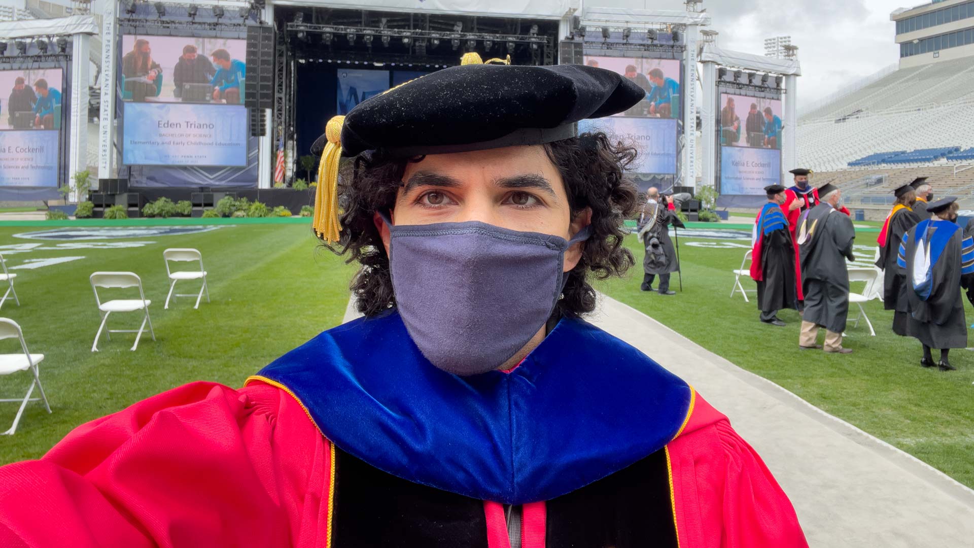 Me in a red University of Maryland PhD robe, from the shoulders up, wearing a black tam and a pandemic mask. In the background, pandemic-spaced folding chairs and a graduation stage; other faculty standing around.