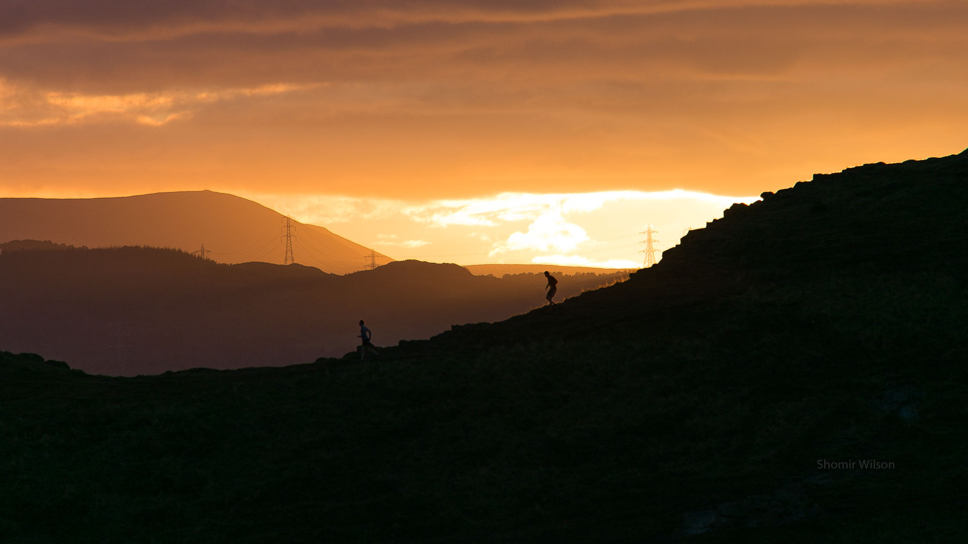 joggers along a ridge line with a bright orange sunset behind them
