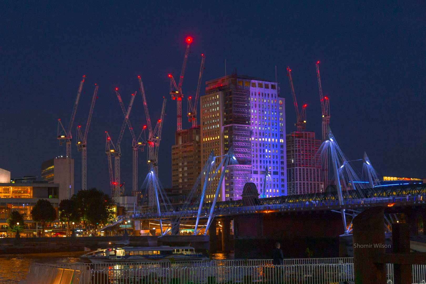 construction cranes around a tall building at night; the cranes have red lights and the building is lit in purple