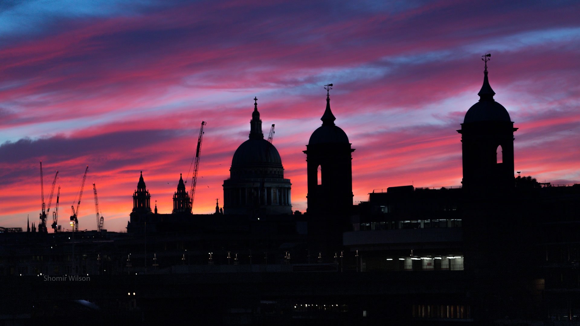 silhouettes of domed buildings and construction cranes against wispy red sunset clouds