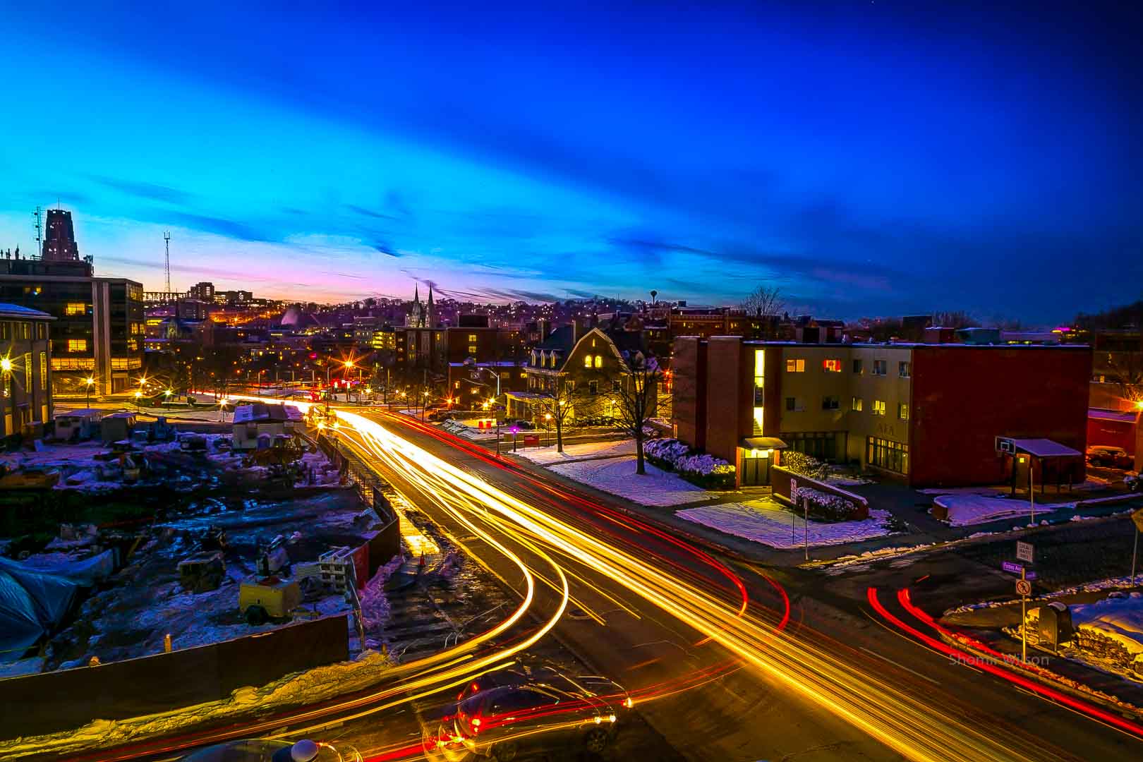 a long exposure of a street intersection in a city at night, showing streaks of headlights and tailights; the sky glows blue