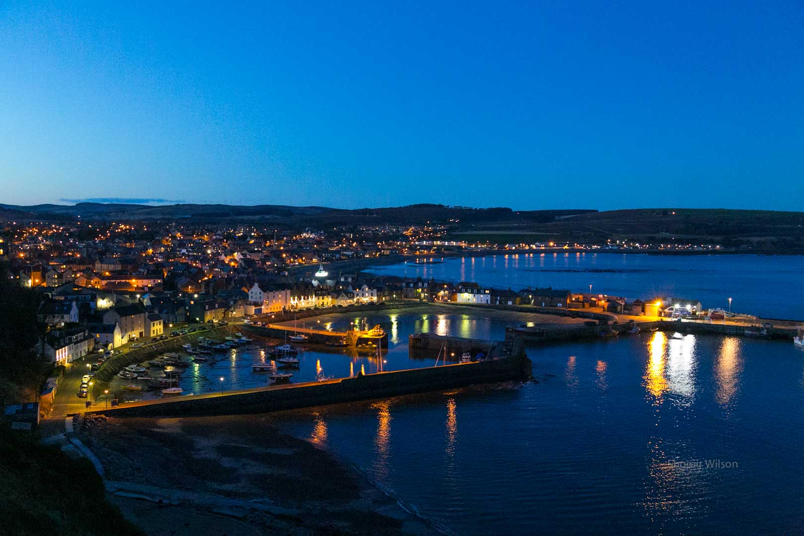 the harbor of a small seaside town at night, taken from a nearby hill