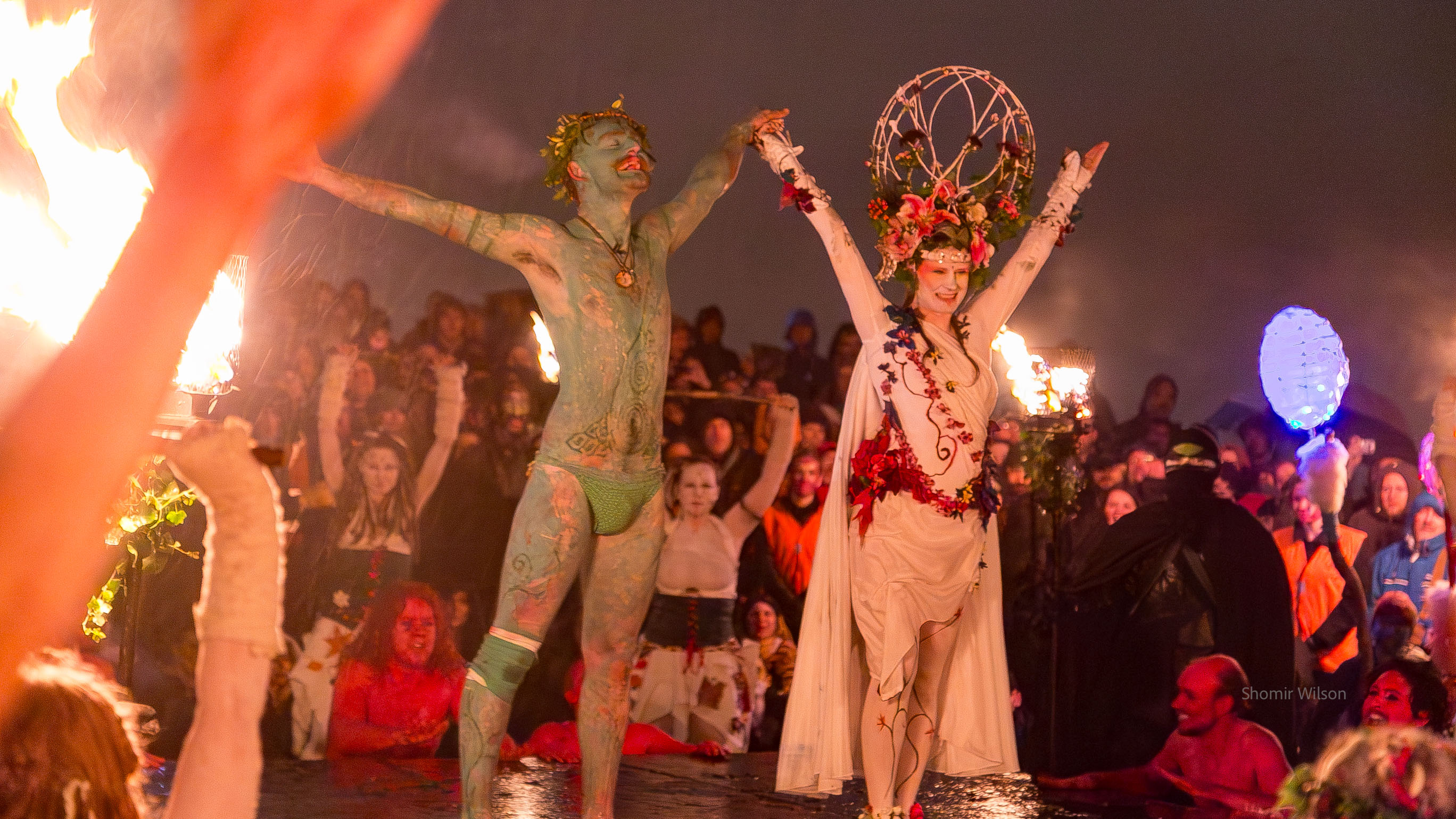 Costumed Green Man and May Queen with other costumed performes in the background, at night outside