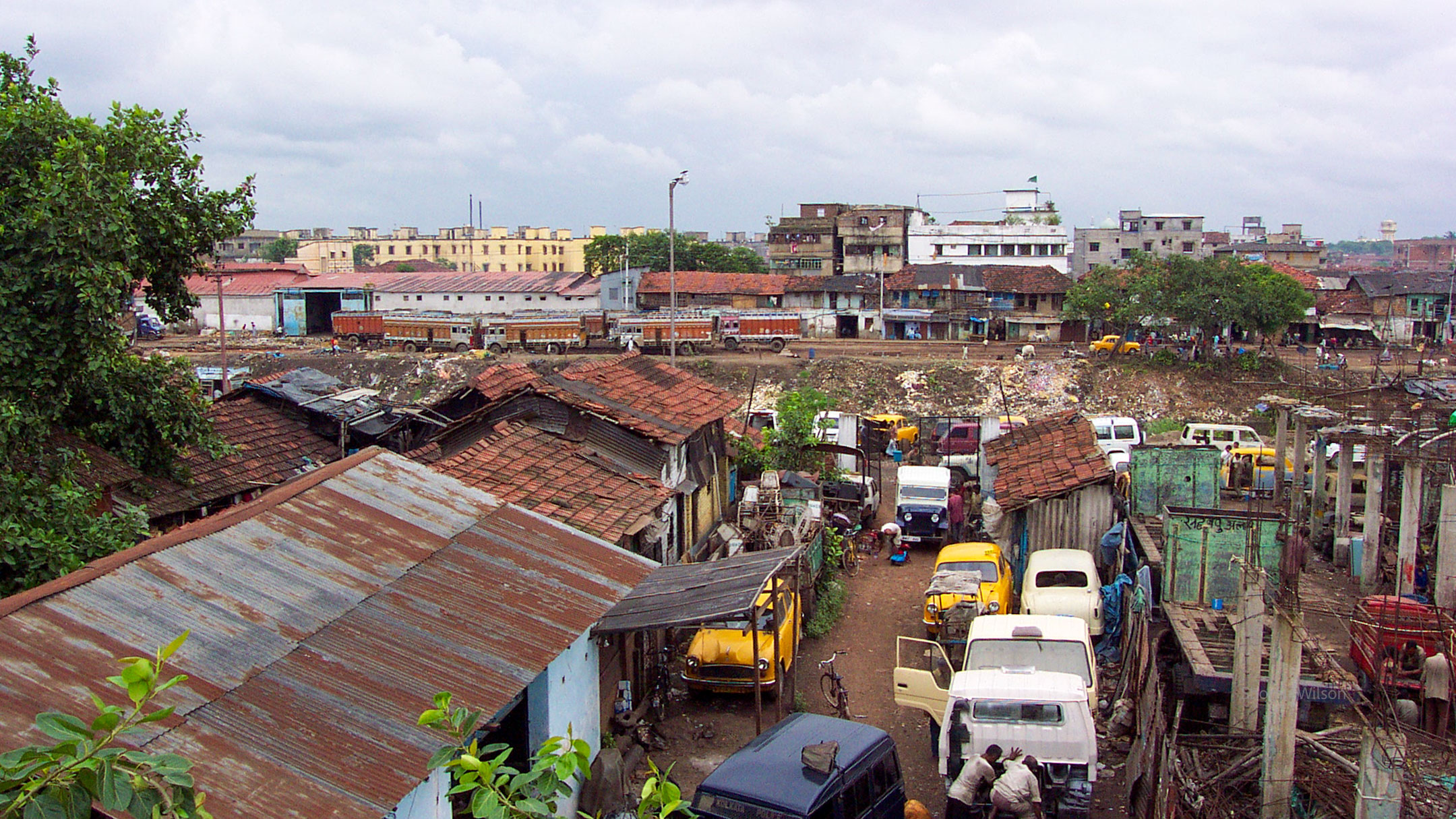 From a height of about 30 feet, a cluttered dirt street with ramshackle buildings and lots of parked cars