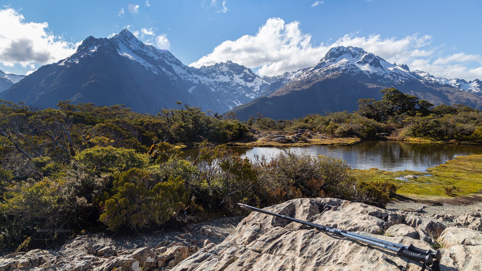 foreground, below: trekking poles, a pond, and scrubby foliage; background, above: mountains and blue sky with a few clouds