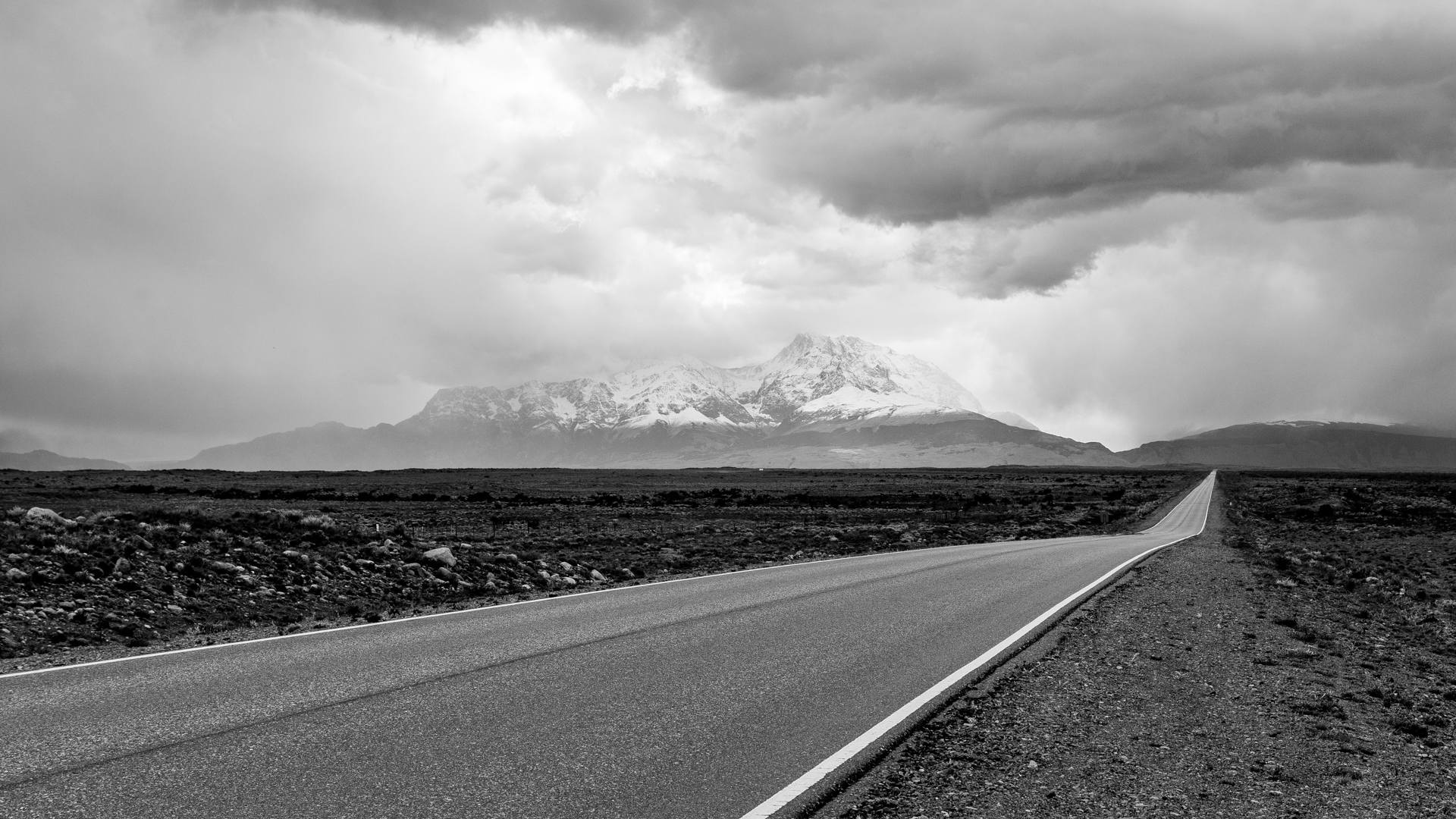 black and white: a two-lane road across a dessert with mountains in the distance and cloudy sky above