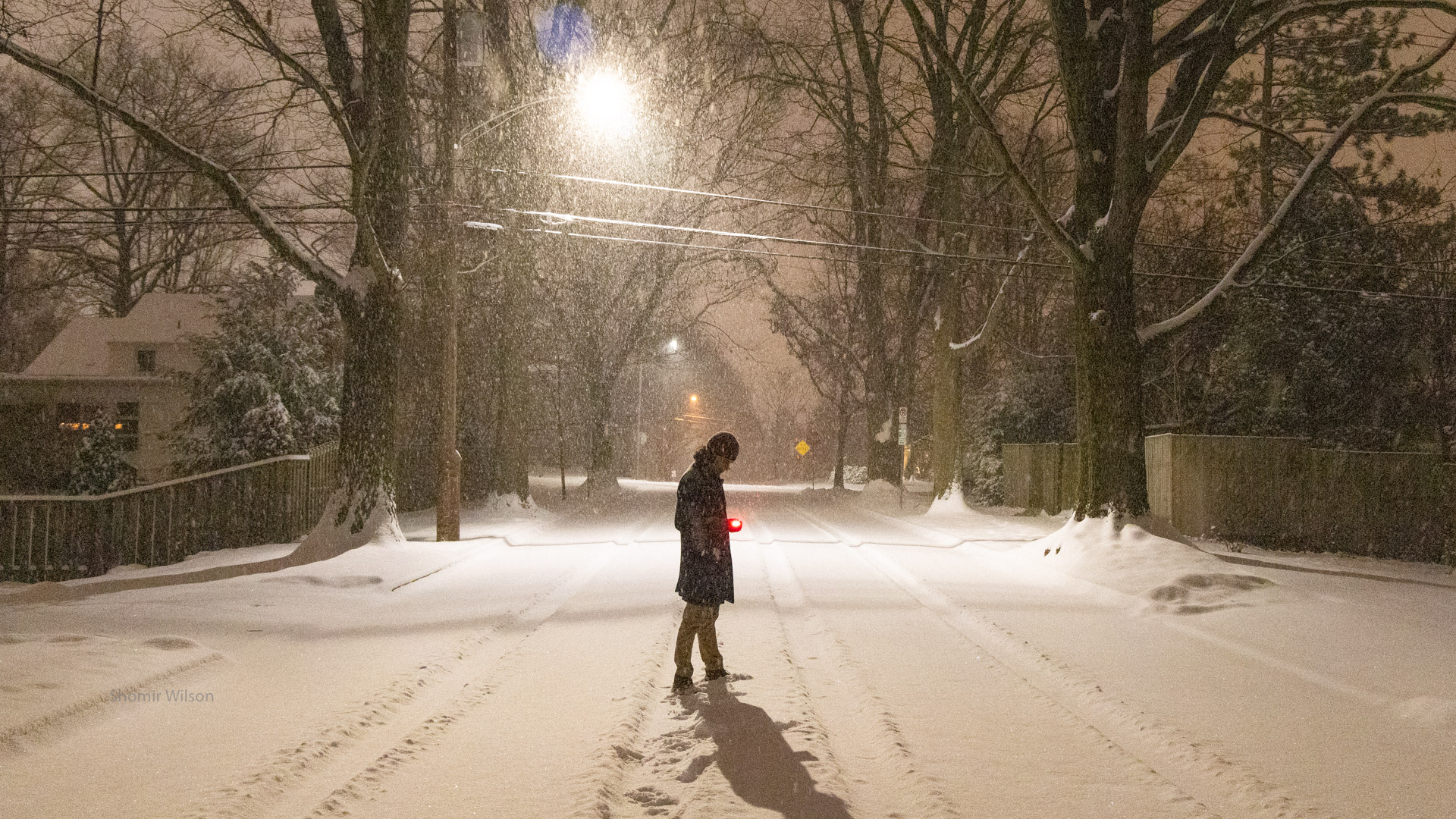 person standing in the middle of a snowy residential neighborhood street at night, standing in profile, holding a mysterious bright red light
