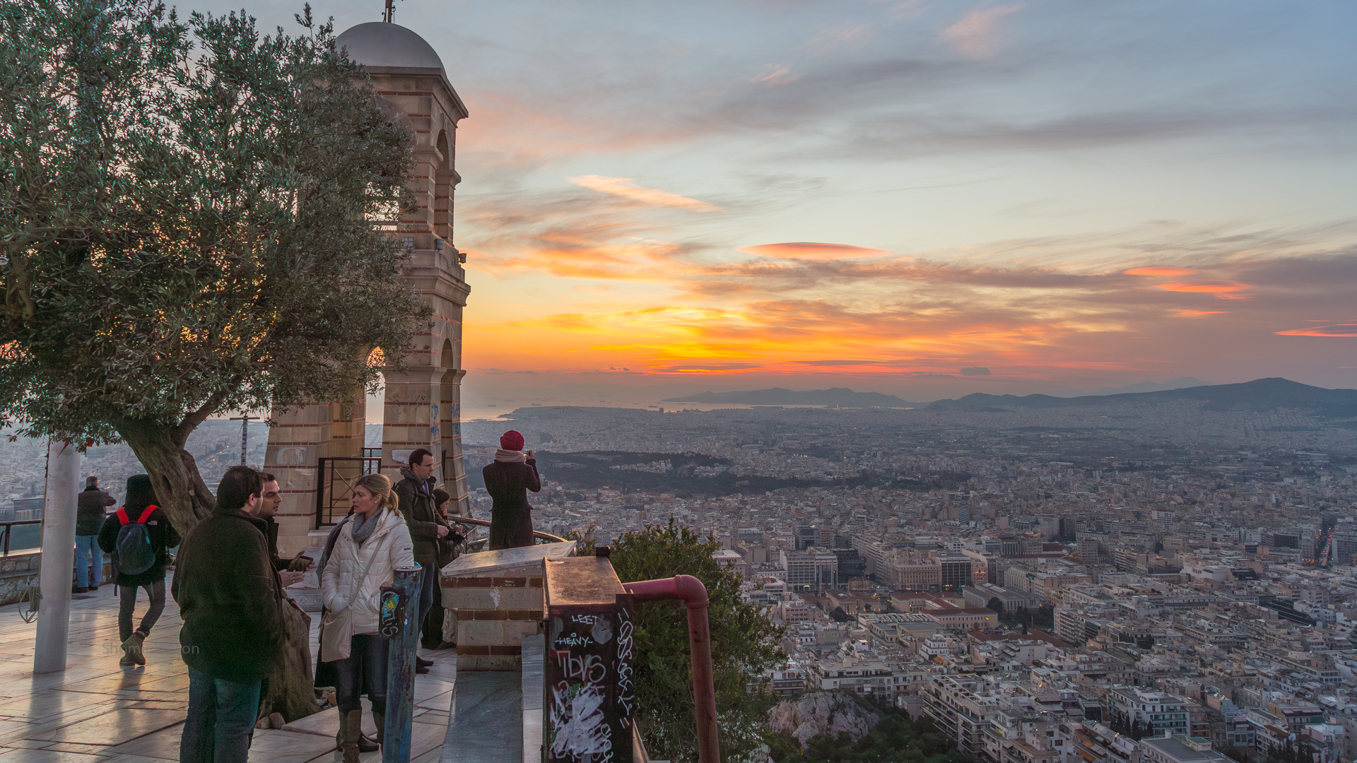 to the left, a stone observation deck with several people, a tree, and a bell tower; to the right, below, a city; the sky has an orange sunset